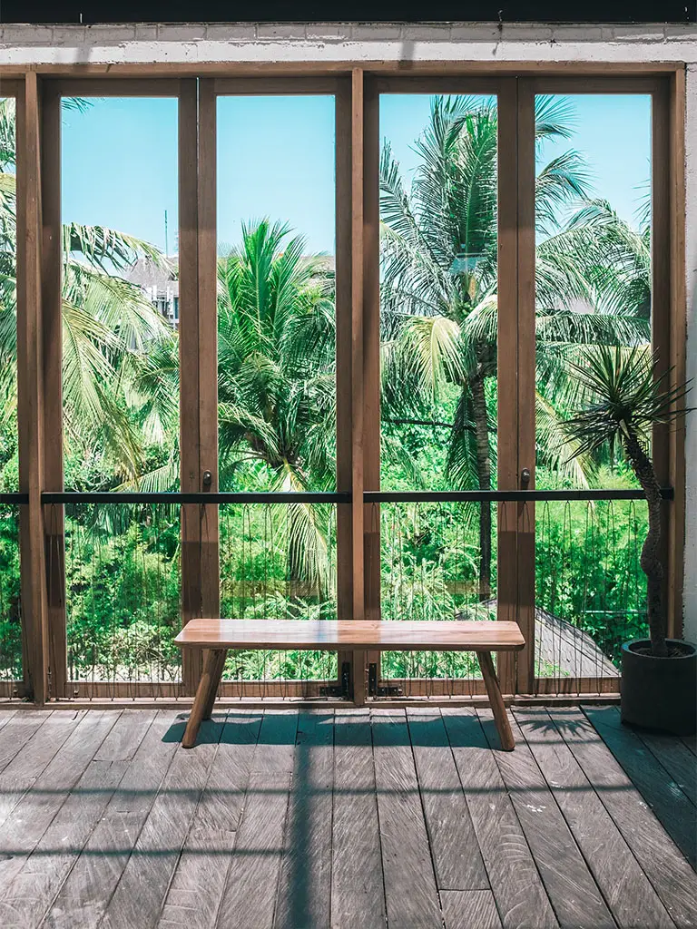 Inside an eco house looking out of a large window at palm trees and garden with a bench in front.