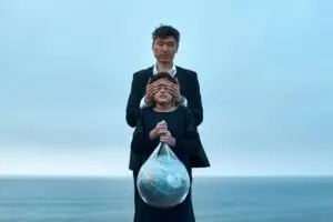 Man holding his hands over a child's eyes while she is holding a bag of trash with a globe inside it. They are in front of the ocean.